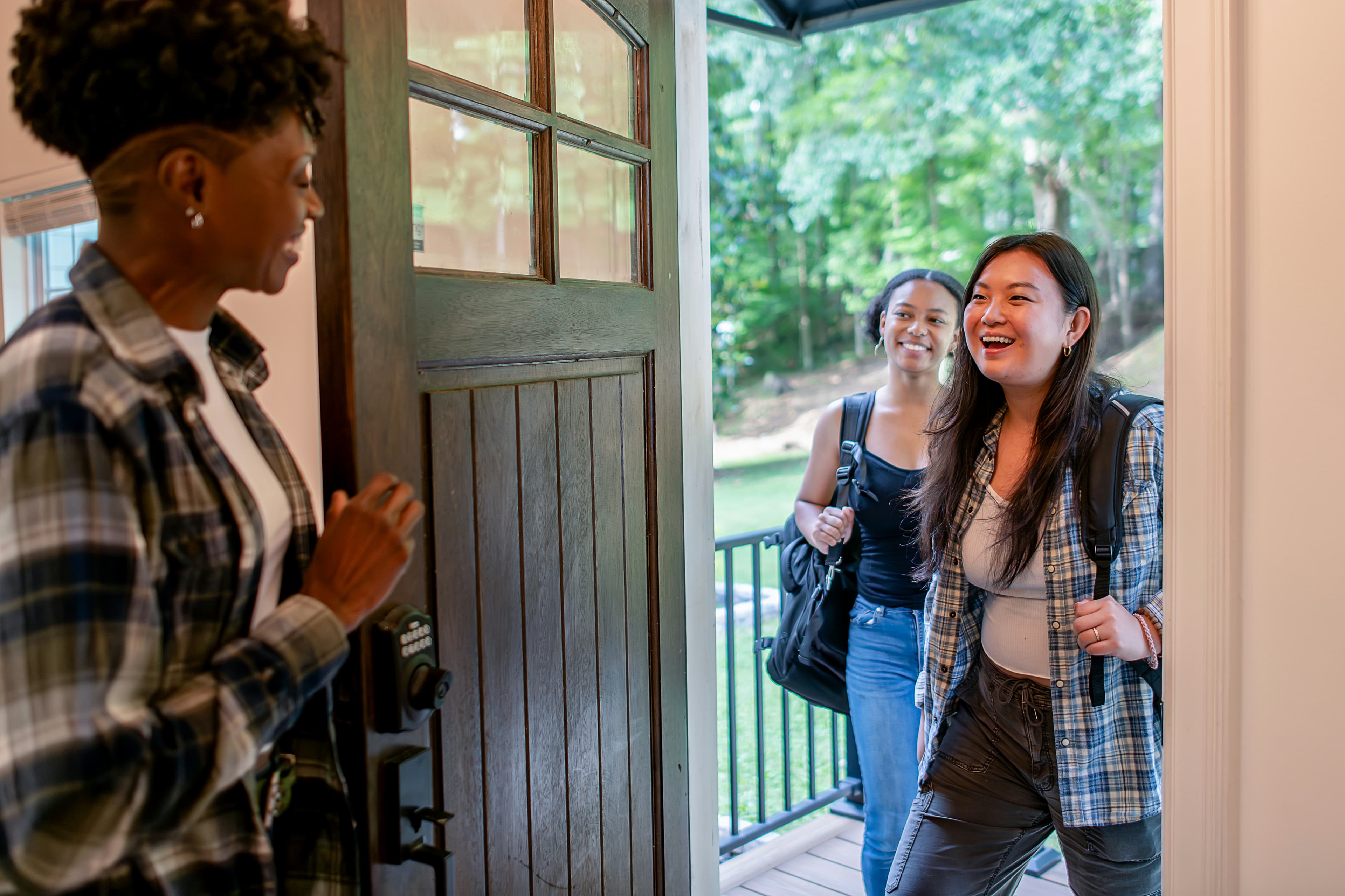 A person holding open a front door for two smiling visitors carrying backpacks
