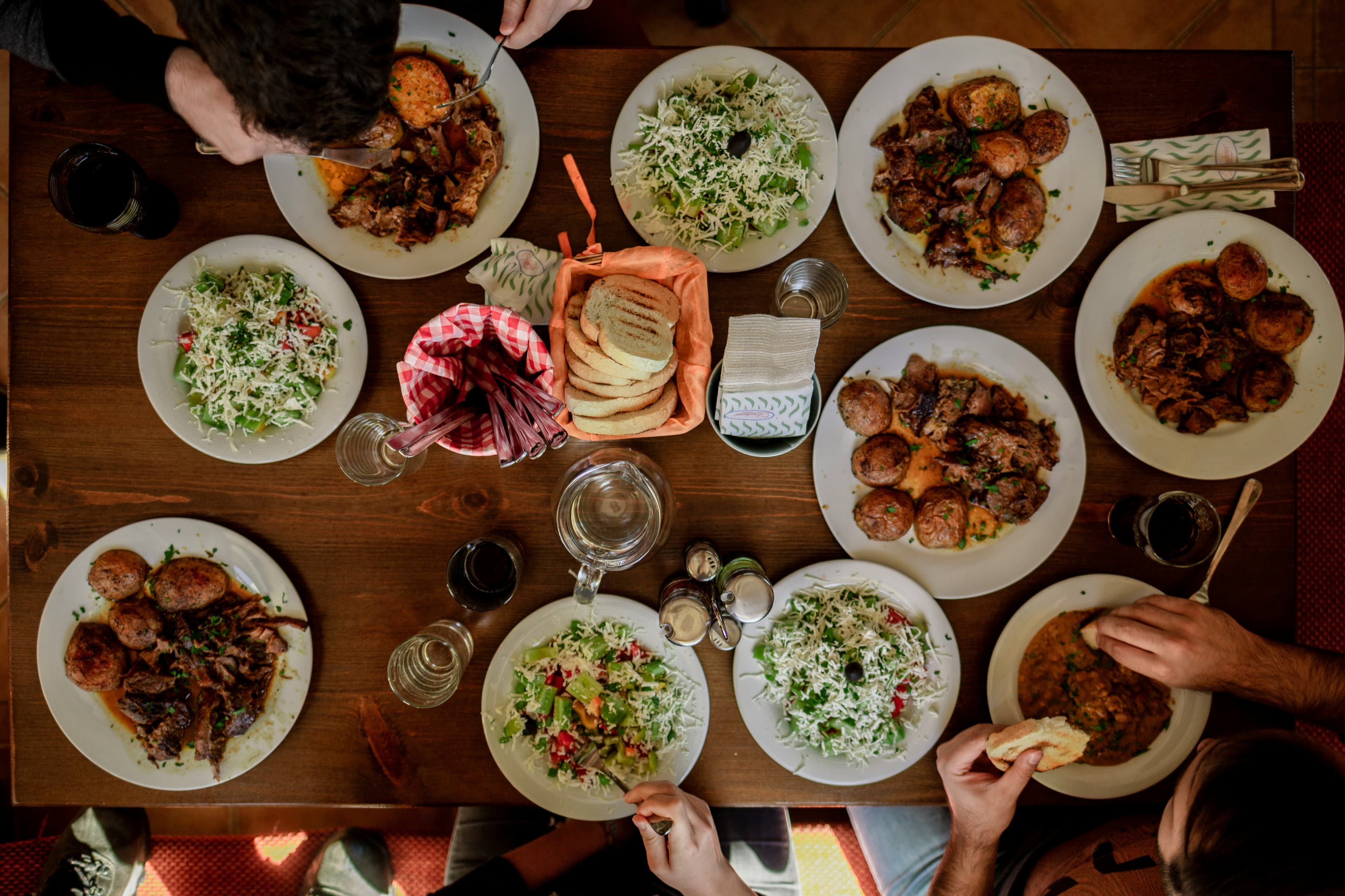 Overhead view of a shared meal on a wooden table, with plates of salad and roasted meat and several people reaching in