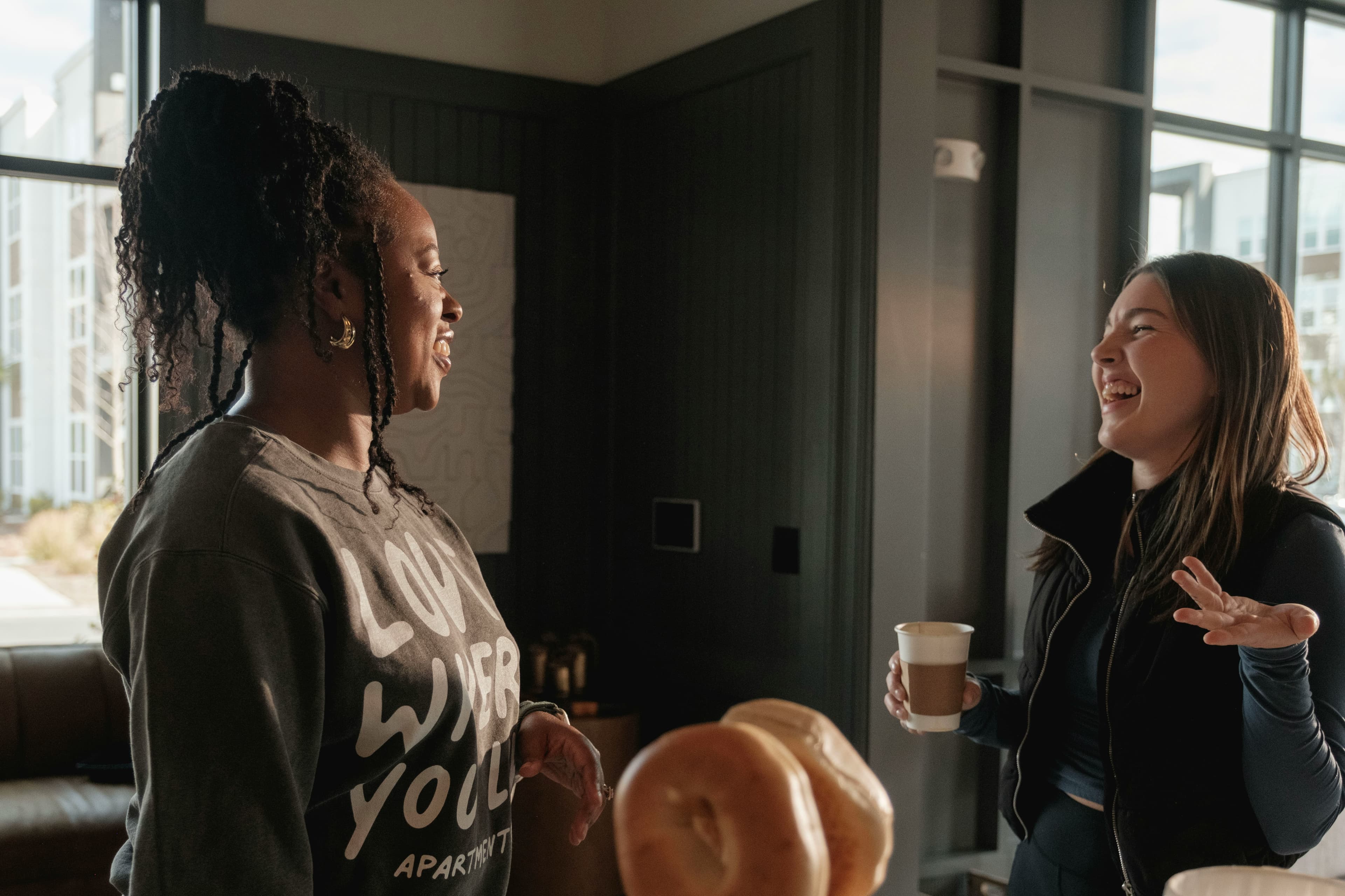 Two people laughing together in a bright room with large windows, one holding a coffee cup