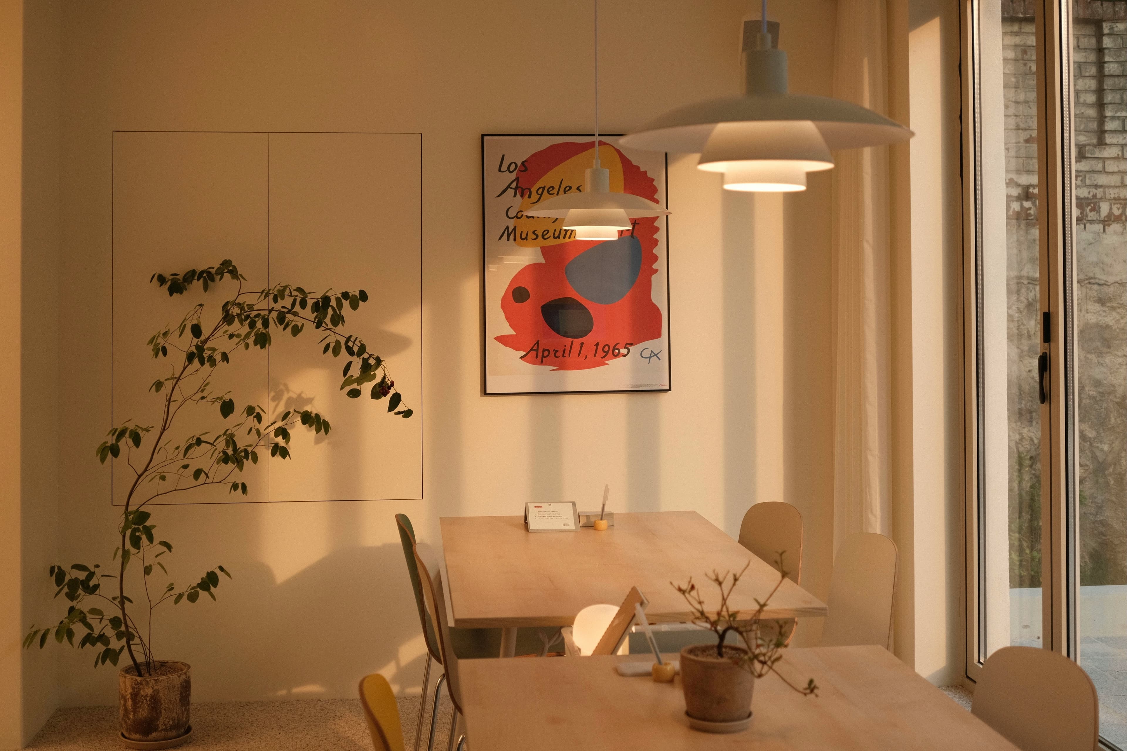 A sunlit room with wooden tables and chairs, pendant lamps, potted plants, and a framed poster on the wall