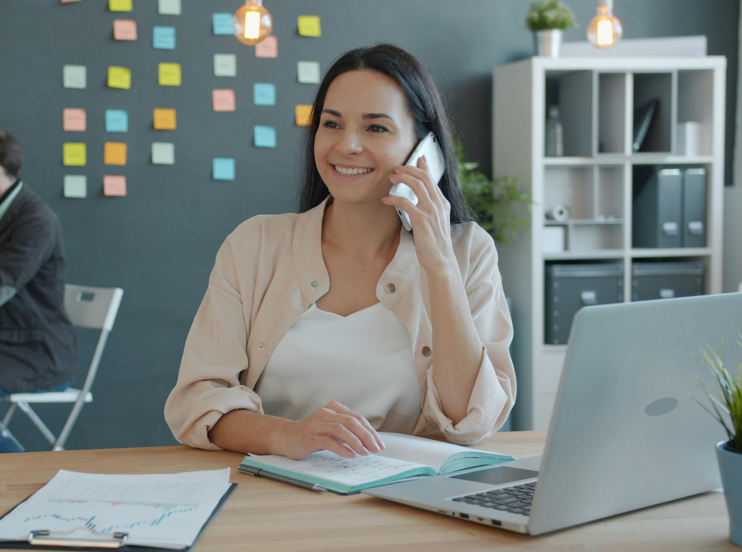 A smiling woman talking on a phone at a desk with an open notebook and a laptop