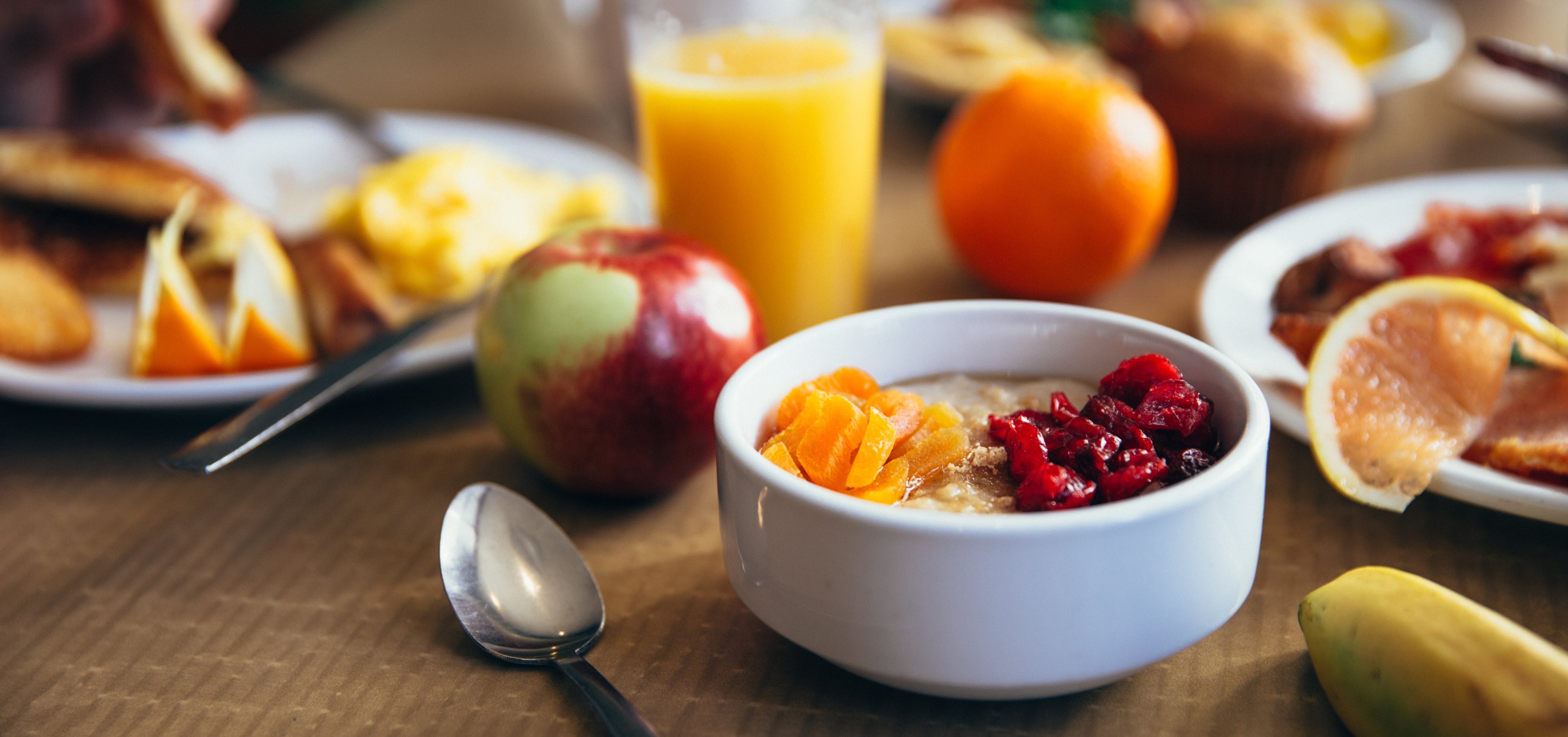 A breakfast table with oatmeal topped with dried fruit, orange juice, an apple, and an orange