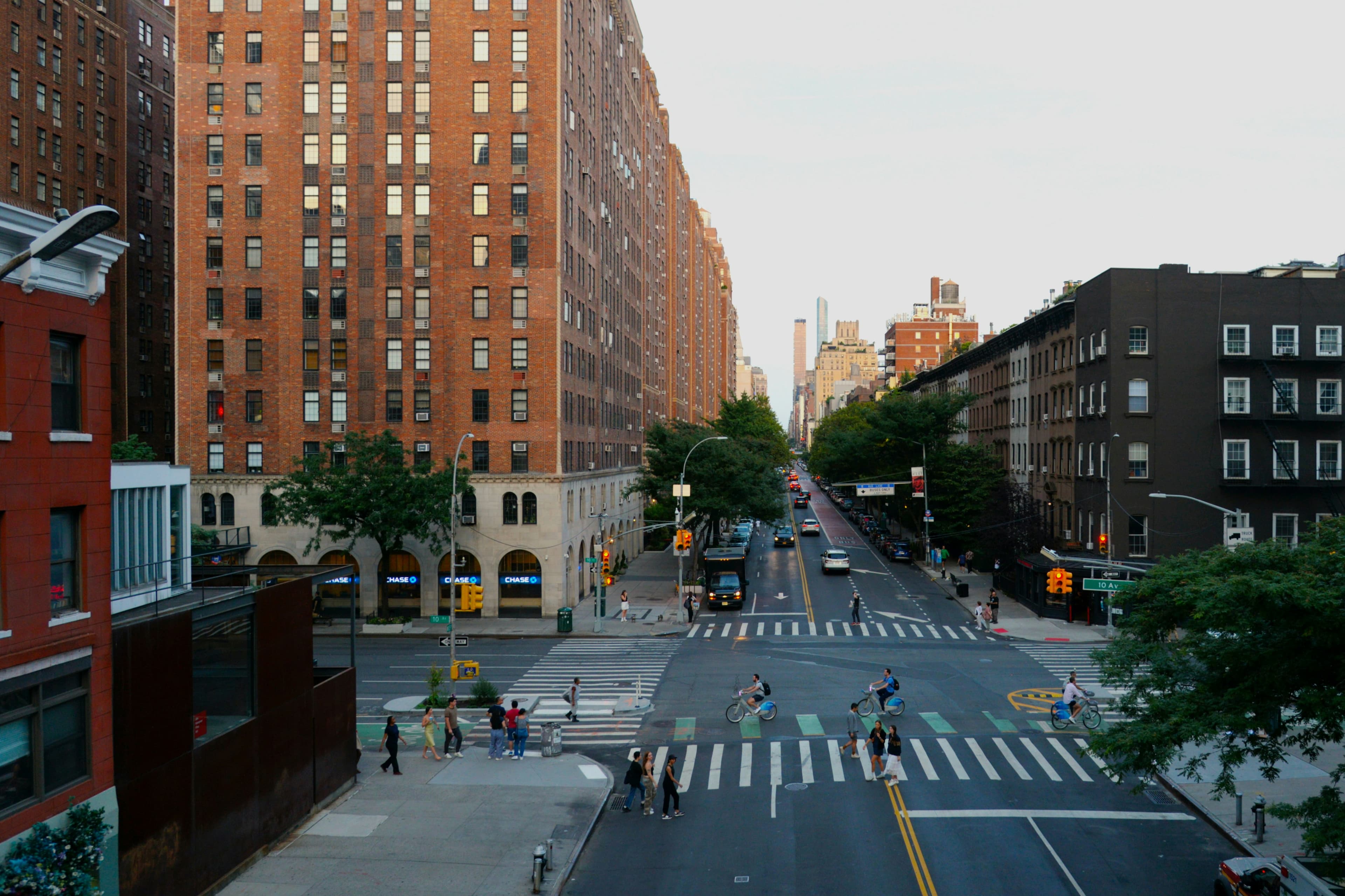 A city street with crosswalks, cyclists, and people walking between tall brick buildings